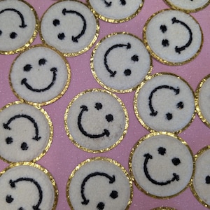 May include: A selection of white, circular patches featuring black smiley faces and gold glitter edging. The patches are arranged on a pink surface, creating a cheerful pattern. The smiley faces have different expressions.