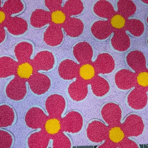 May include: A close-up of a textile featuring a repeating floral design. The flowers are a bright pink with yellow centres and light blue outlines. The background is a light purple, creating a cheerful and playful pattern.