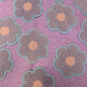 May include: Close-up of several flower-shaped patches. Each patch has a grey, textured petal design with a light peach centre and a light blue border. The patches are arranged on a purple fabric background.