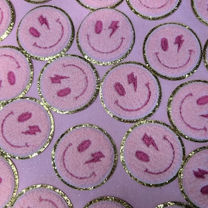 May include: A collection of pink and gold smiley face patches with a lightning bolt design. The patches are made of a soft, fuzzy material and have a gold glitter trim.