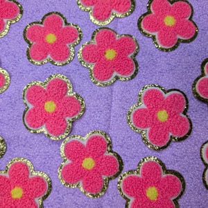 May include: Pink flower-shaped patches with yellow centres and gold glitter borders are scattered across a purple fabric background. The patches have a textured, fuzzy appearance, and are evenly spaced.