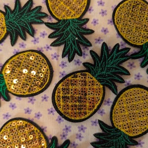 May include: Embroidered pineapple patches with gold sequins and green leaves. The patches are outlined in black and are arranged on a white fabric with small purple flower patterns.