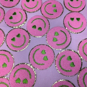 May include: A collection of pink and green smiley face patches with a gold glitter outline. The smiley faces have green hearts for eyes and a green smile.
