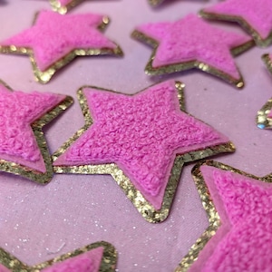 May include: Pink fuzzy star-shaped patches with gold trim. The stars are scattered on a pink sparkly background.
