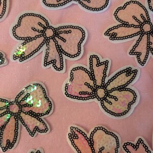 Könnte beinhalten: Rosafarbener Stoff mit mehreren Paillettenbögen und -blumen. Die Bögen und Blumen sind schwarz umrandet und haben einen weißen Hintergrund.