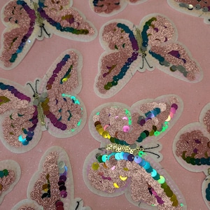 May include: A collection of pink butterfly-shaped fabric patches with iridescent sequins. The patches are on a pink background.