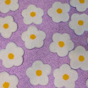 May include: White, flower-shaped appliques with yellow centres are scattered across a textured, lavender surface. The appliques have a soft, fuzzy texture, suggesting a craft or decorative theme.