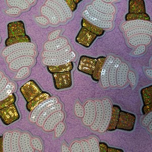 Könnte beinhalten: Eine Sammlung von paillettenbesetzten Eiscreme-Kegel-Patches. Das Eis ist weiß mit rosa Besatz, und die Kegel sind goldfarben. Die Patches sind auf einem lila Hintergrund verstreut.