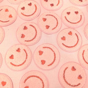May include: A pink background with multiple pink fuzzy smiley faces with red embroidered hearts for eyes.