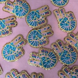 May include: A collection of embroidered iron-on patches featuring a peace sign with an eye in the center. The patches are pink, blue, and gold with glitter accents.