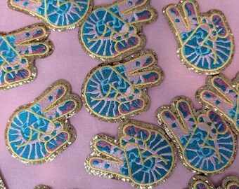 Könnte beinhalten: Eine Sammlung von gestickten Bügeletiketten mit einem Peace-Zeichen mit einem Auge in der Mitte. Die Patches sind rosa, blau und goldfarben mit Glitzerakzenten.