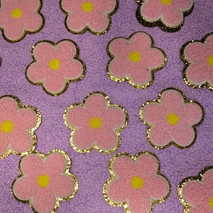 May include: Pink flower-shaped patches with yellow centres and gold glitter edging are arranged on a purple textured surface. These decorative patches are ideal for adding a floral touch to clothing, accessories, or crafts.