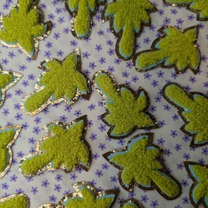 Puede incluir: Primer plano de varios parches de tela verdes con forma de hoja y aspecto texturizado. Cada parche tiene un borde de purpurina dorada y un contorno azul claro, sobre una tela blanca con pequeños estampados florales morados.