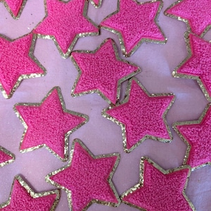 May include: A collection of pink fuzzy star-shaped patches with gold glitter trim. The patches are arranged on a pink background.