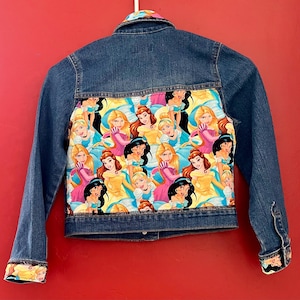 May include: A denim jacket with a colorful princess-themed fabric panel on the back. The jacket features a classic collar, button closures, and cuffs with princess-themed fabric. The jacket is a medium wash blue.