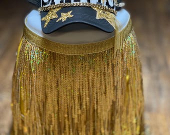 May include: A black and white trucker hat with gold chain accents and the word "SAINT" embroidered on the front. The hat is sitting on top of a gold fringe skirt.