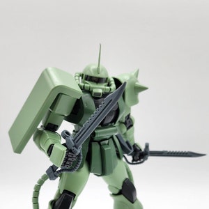 May include: A green and black plastic action figure of a robot with a large shield and two swords. The robot has a detailed design and is posed in a fighting stance.