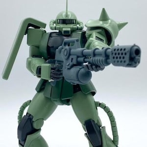 以下が含まれることがあります： 銃を持ったロボットの緑と黒のプラスチック製アクションフィギュア。ロボットは、バイザーと大きな胸部を持つ大きな頭を持っています。ロボットは白い表面に立っています。