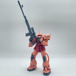 Könnte beinhalten: Eine rote und graue Plastik-Actionfigur eines Roboters, der ein schwarzes Gewehr hält. Der Roboter hat einen roten Helm und einen roten und grauen Körper. Der Roboter steht auf einer weißen Oberfläche.