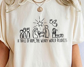 May include: Cream-coloured t-shirt with a black line drawing of a nativity scene, a star, an angel, and the text "A thrill of hope, the weary world rejoices." The shirt has short sleeves and a relaxed fit.