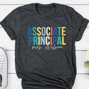 May include: A dark gray t-shirt with the text "ASSOCIATE PRINCIPAL" in colorful letters and "mrs. Wilson" in white script below.