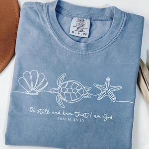 May include: A blue Comfort Colors t-shirt featuring a white line drawing of a seashell, turtle, and starfish. The text "Be still and know that I am God, Psalm 46:10" is printed below the design. The shirt is a size S-P-CH.