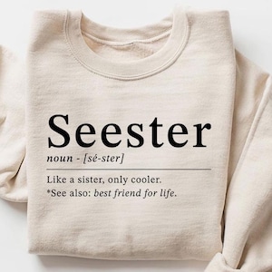 May include: A beige crewneck sweatshirt with the word "Seester" printed in black, along with a definition. The definition reads "Like a sister, only cooler." and "See also: best friend for life."