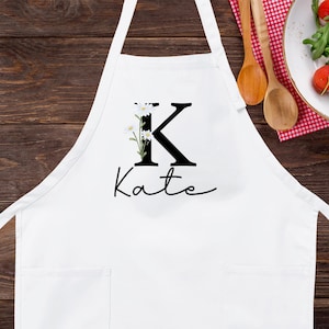 May include: White kitchen apron with a black letter K and the name Kate printed on it. The letter K is decorated with small white flowers.