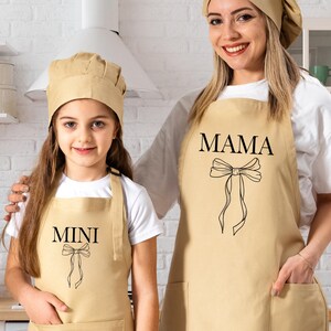 May include: Two beige aprons with a bow design. The larger apron says "MAMA" and the smaller apron says "MINI".