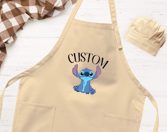 Puede incluir: Un delantal de color beige con un gráfico de un personaje alienígena azul de orejas grandes y la palabra CUSTOM impresa en negro encima. El delantal incluye dos bolsillos delanteros y se acompaña de un gorro de chef beige a juego con pliegues.