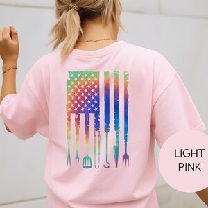 May include: Light pink t-shirt with a colorful graphic of the American flag and grilling tools. The flag design features a rainbow gradient and stars, with grilling utensils replacing the stripes. The shirt has short sleeves.