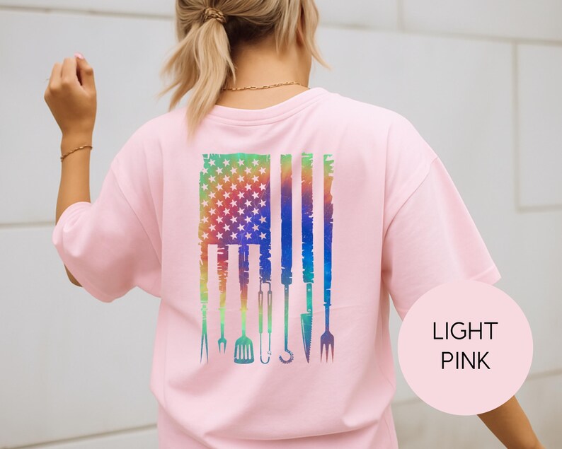 May include: Light pink t-shirt with a colorful graphic of the American flag and grilling tools. The flag design features a rainbow gradient and stars, with grilling utensils replacing the stripes. The shirt has short sleeves.