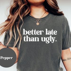 May include: Pepper-colored t-shirt with the phrase "better late than ugly" in white block letters. The shirt has a crew neck and short sleeves. The image also shows a person wearing a necklace and bracelet.