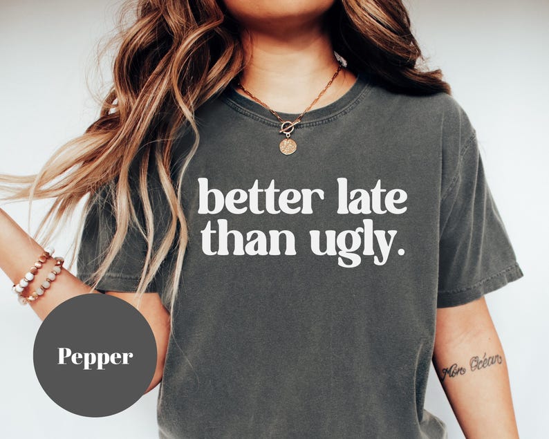 May include: Pepper-colored t-shirt with the phrase "better late than ugly" in white block letters. The shirt has a crew neck and short sleeves. The image also shows a person wearing a necklace and bracelet.