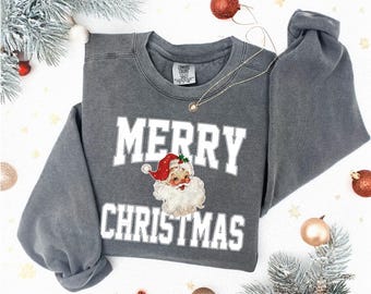 May include: A grey sweatshirt featuring the text "MERRY CHRISTMAS" in white, with a vintage Santa Claus illustration. The image is set against a backdrop of Christmas decorations, including baubles and foliage.