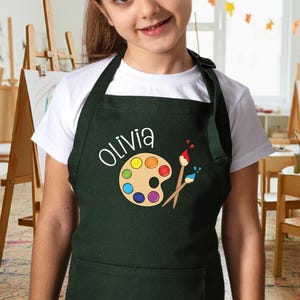 May include: A green child's apron with a paint palette and paintbrushes design. The apron has a pocket and the name "Olivia" is embroidered on the front.