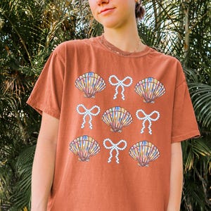 Comfort Colors t-shirt with a seashell and bow design, perfect for beach days, coastal vibes, and summer outings with a preppy aesthetic