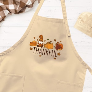 May include: A beige apron with two front pockets and a matching chef hat. The apron features a graphic print of a slice of pumpkin pie, a gnome, a pumpkin, and a turkey, with the text THANKFUL and scattered autumn leaves in brown and orange.