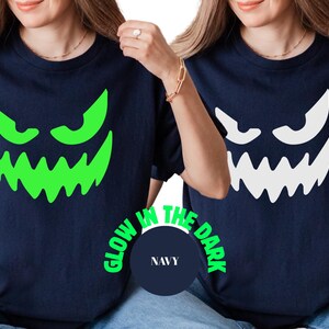 May include: Navy blue t-shirts featuring a glowing green and white jack-o'-lantern face design. The text "GLOW IN THE DARK" is printed around a navy blue circle with the word "NAVY" inside. The shirts are Halloween themed.