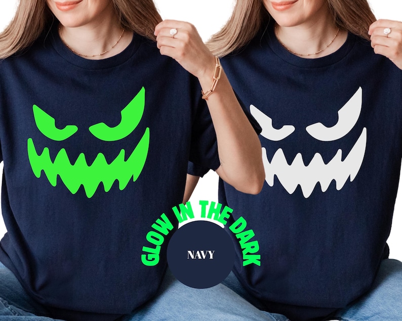 May include: Navy blue t-shirts featuring a glowing green and white jack-o'-lantern face design. The text "GLOW IN THE DARK" is printed around a navy blue circle with the word "NAVY" inside. The shirts are Halloween themed.