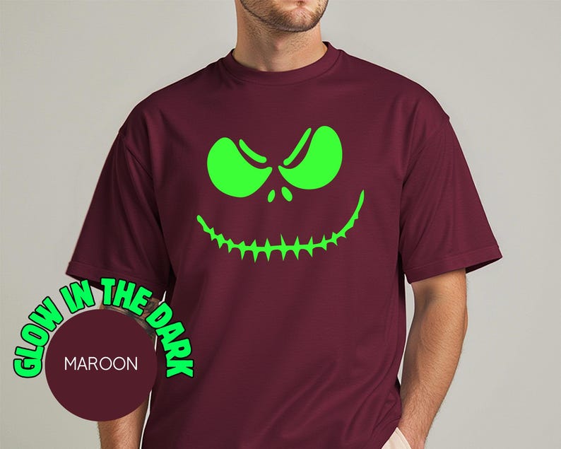 May include: Maroon t-shirt with a glow-in-the-dark green face design. The design features a grinning face with sharp teeth and angry eyes. The text "GLOW IN THE DARK" is printed around a maroon circle with the word "MAROON" inside.