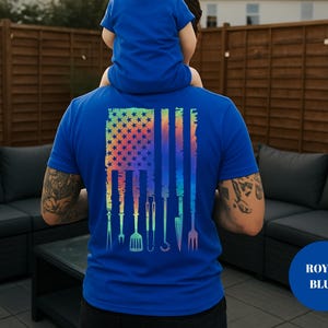 May include: Royal blue t-shirt featuring a colorful American flag design with grilling tools. The flag design includes stars and stripes, with various barbecue utensils replacing the stripes. A child is sitting on the person's shoulders.