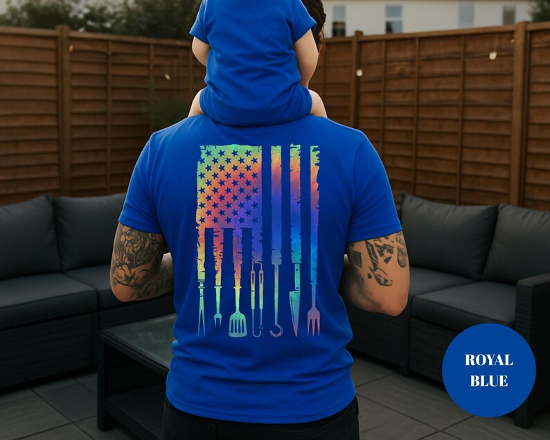 May include: Royal blue t-shirt featuring a colorful American flag design with grilling tools. The flag design includes stars and stripes, with various barbecue utensils replacing the stripes. A child is sitting on the person's shoulders.