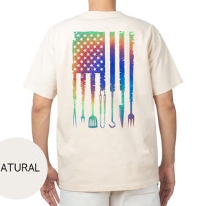 May include: A natural-colored t-shirt featuring a distressed American flag design on the back. The flag is rendered in a rainbow gradient, with grilling tools replacing the stripes. The word "NATURAL" is printed on a circular label.