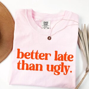 May include: A light pink t-shirt with the phrase "better late than ugly" in orange block letters. A gold necklace with a small pendant is draped on the shirt. A brown hat and dried palm leaf are in the background.