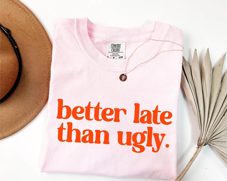 May include: A light pink t-shirt with the phrase "better late than ugly" in orange block letters. A gold necklace with a small pendant is draped on the shirt. A brown hat and dried palm leaf are in the background.