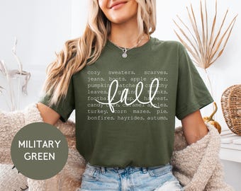 May include: Military green t-shirt with the word "fall" in a cursive font. The shirt lists fall-themed words like "sweaters," "pumpkin patches," and "hayrides" in a white font. The shirt is being worn by a person.