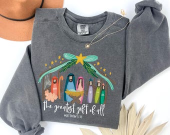 May include: Grey crew neck sweatshirt with a colourful Nativity scene illustration. The design includes a stable, figures, and the text "the greatest gift of all" with Matthew 1:21 below. A brown hat and gold necklace are also visible.