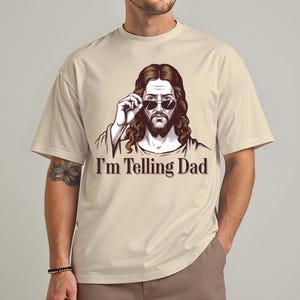 May include: A beige t-shirt with a graphic of Jesus wearing sunglasses and the text "I'm Telling Dad".