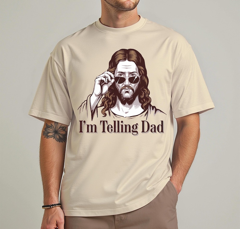 May include: A beige t-shirt with a graphic of Jesus wearing sunglasses and the text "I'm Telling Dad".